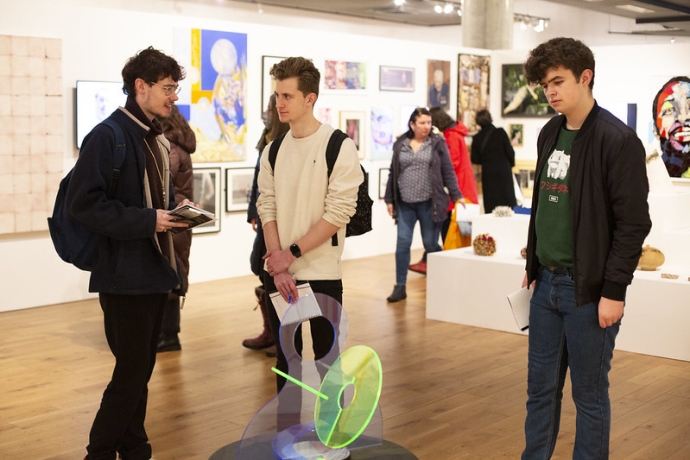 Three young males stand discussing a sculpture on the floor in a gallery 
