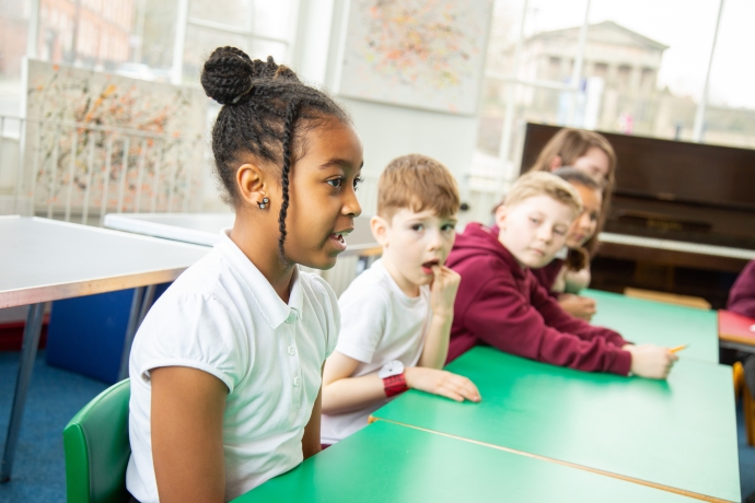 A young black female student in the forefront talking to someone off camera, sat at a desk. Three white male and female students are sitting next to her at the table