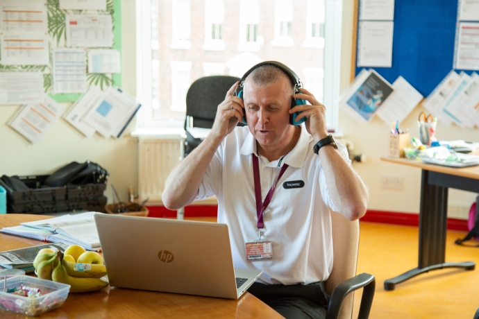 A white male teacher in a white t-shirt sitting in front of a laptop putting ear defenders on