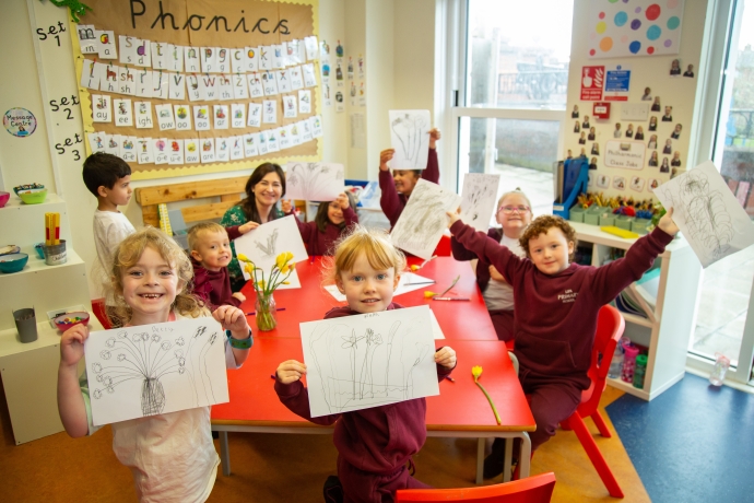 Children in a classroom standing up around the table showing off their drawings