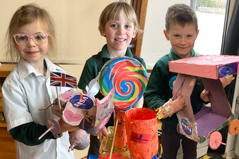 Three children stand side by side, each holding a colorful craft project. The child on the left holds a model featuring a Union Jack flag and NASA logo. The middle child displays a large, multicolored lollipop decoration. The child on the right holds a pink and purple model resembling a vehicle or structure. 