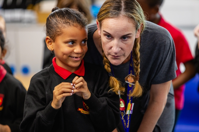 A young student wearing a black school uniform with a red collar is standing next to an adult with braided hair, who is leaning in close. Both of their faces are blurred for privacy. The background shows other students and classroom elements out of focus
