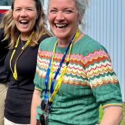 A woman with grey hair and a bright green top is looking to the camera, smiling. Another woman is smiling behind her.